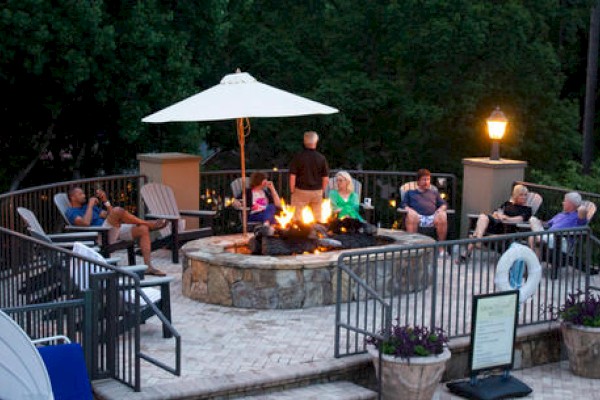 A group of people are sitting around a fire pit on a patio, surrounded by trees, with an umbrella and lit lamps creating a cozy ambiance.