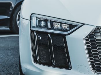 The image shows the front view of a white sports car, parked closely to another vehicle, showcasing its headlight and grille.