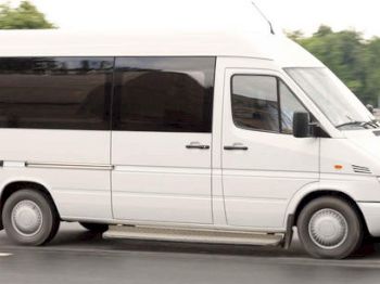 The image shows a white van driving on a road, with trees and a fence visible in the background. The van has tinted windows on the side.