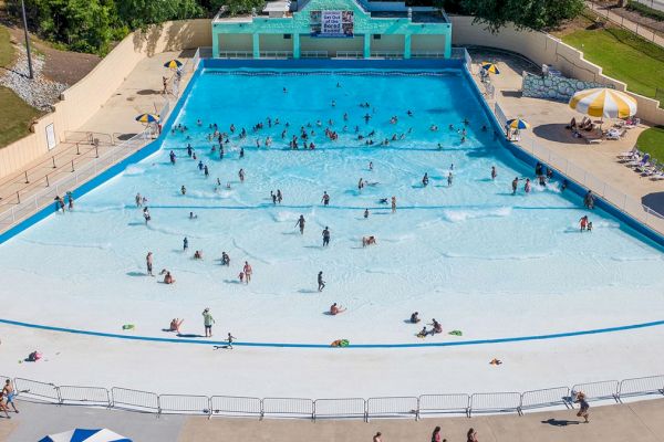 The image shows a wave pool with people enjoying the water. There are lounge chairs and tables with umbrellas around the pool area.