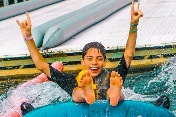 A young boy is enjoying a ride down a water slide in an inflatable tube, throwing up a hand gesture and smiling, with water splashing around.
