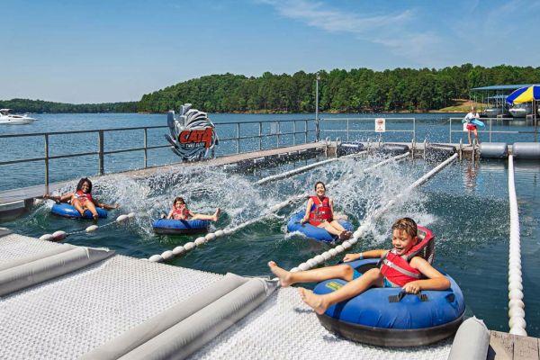 Four people are riding on water tubes, splashing down a water slide into a lake, set against a backdrop of trees and a docked boat.