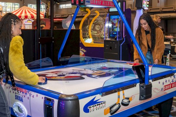 Two people are playing air hockey in an arcade filled with various games and attractions.
