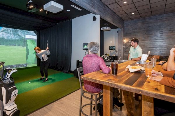 Four people are in a room, with one person playing virtual golf while the others watch and enjoy drinks at a wooden table.