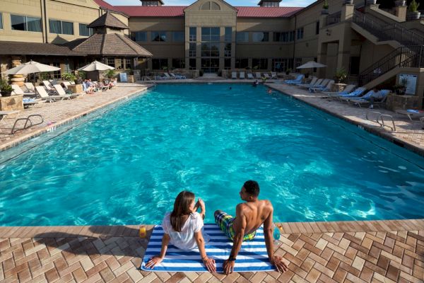 The image shows two people sitting at the edge of a large outdoor pool at a resort, with the buildings and pool chairs surrounding the pool.