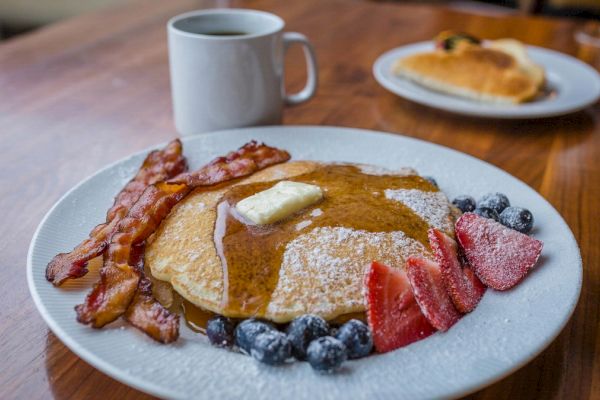 The image shows a breakfast plate with pancakes topped with butter and syrup, bacon, blueberries, and strawberries, next to a mug of coffee.