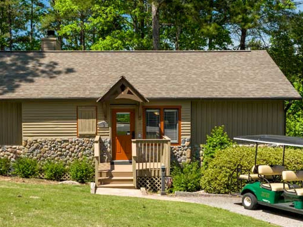 A small, single-story cabin with a porch is surrounded by greenery and has a golf cart parked nearby. The scene appears serene and well-kept.