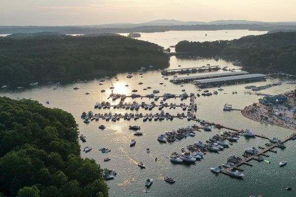 An aerial view of a marina with numerous boats docked, surrounded by lush greenery and calm waters during sunset in the background.