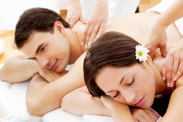 Two people are lying on massage tables, receiving back massages from masseuses. One person has a flower in their hair.