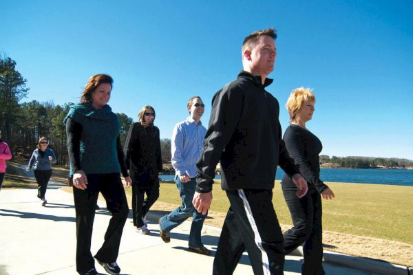 A group of people walking on a paved path near a body of water, under a clear blue sky, with trees in the background.