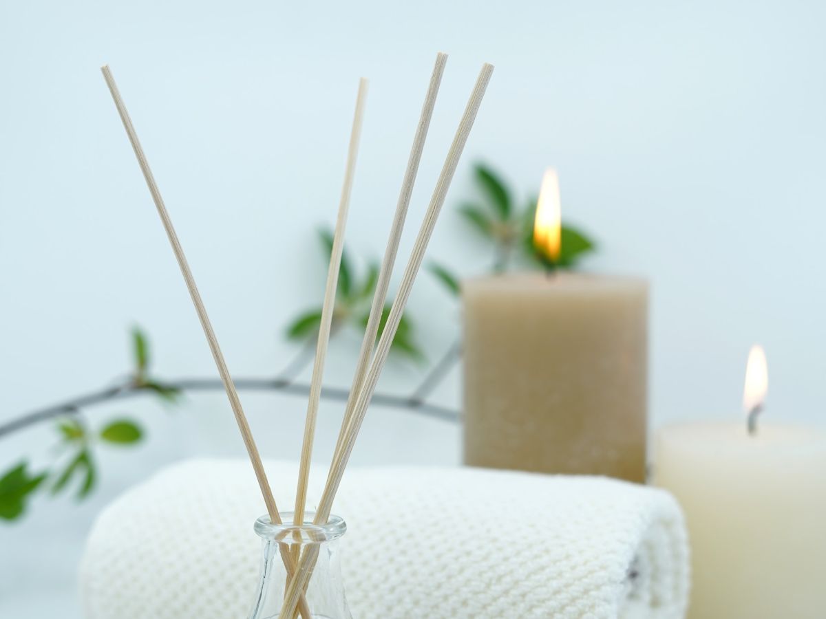 The image shows a spa setup with scented reed diffusers, rolled white towels, and lit candles, accompanied by green leaves.