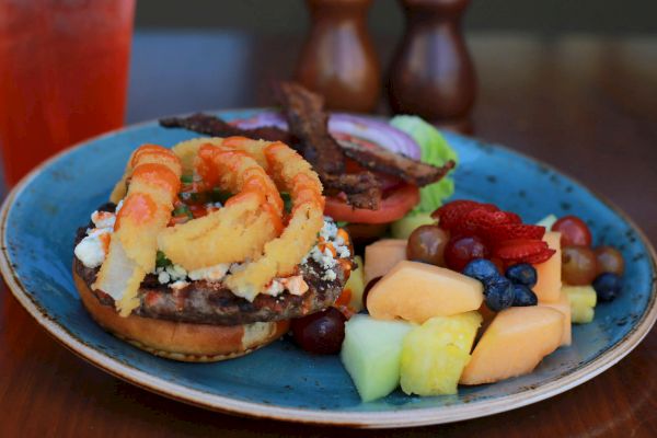 A plate with a burger topped with onion rings and bacon, alongside a serving of mixed fruit, including strawberries, blueberries, melon, and grapes.