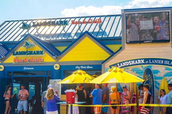 People are waiting outside the Landshark Bar & Grill, a colorful building with outdoor seating, yellow umbrellas, surfboards, and an outdoor screen.