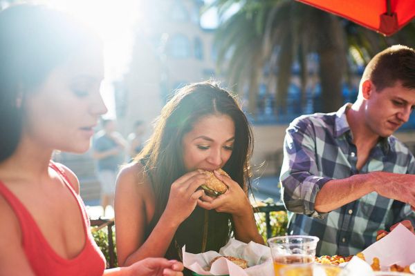 Three people are dining outdoors, with one person eating a hamburger, another person in a red top, and a third person looking at their food.