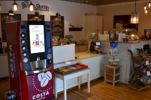 The image shows the interior of a coffee shop, featuring a Costa Coffee vending machine, snacks, a counter with a cash register, and various decorations.