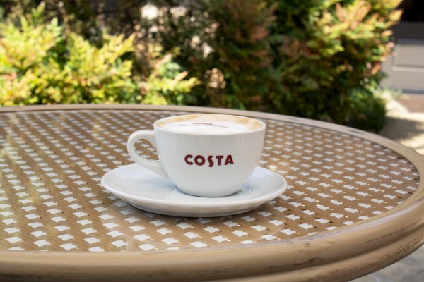 A cup of coffee with foam in a Costa-branded cup on a patterned outdoor table, surrounded by greenery, is pictured here.