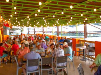 People are dining on an outdoor patio with string lights, colorful decor, and a waterfront view, creating a lively and inviting atmosphere.