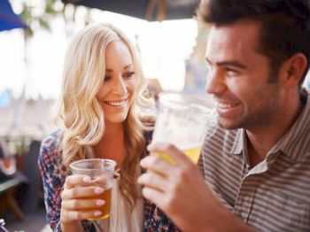 The image shows a woman and a man smiling and holding drinks, seemingly enjoying a pleasant moment together outdoors.