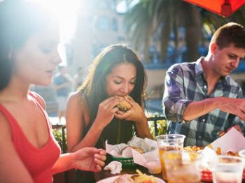 Three people are dining outside, with one person eating a burger. The table is filled with food and drinks, and they seem to be enjoying a sunny day.