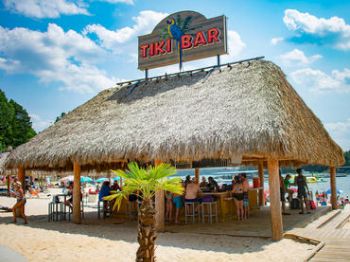 An image shows a tiki bar on a sandy beach, with thatched roof, wooden posts, and people sitting at the bar. The sky is blue and scattered with clouds.