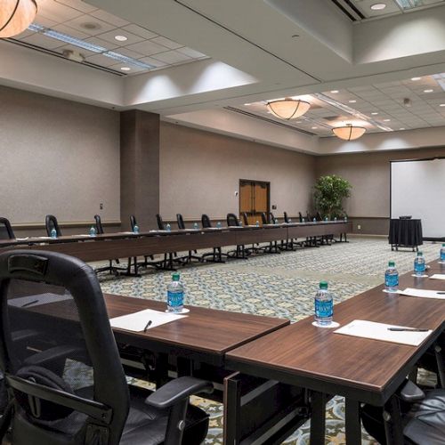 The image shows a large, empty conference room with U-shaped tables, chairs, water bottles, and a projection screen in the background.
