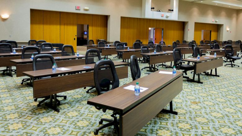 An empty conference room with long tables, office chairs, bottled water, and paper on the tables. The room has a carpeted floor and closed doors.