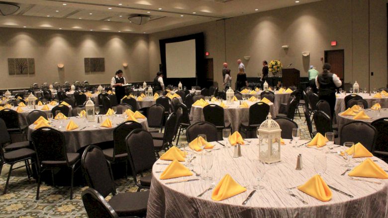 A banquet hall is set up with round tables covered with gray tablecloths and yellow napkins. Some people can be seen arranging items at the back.