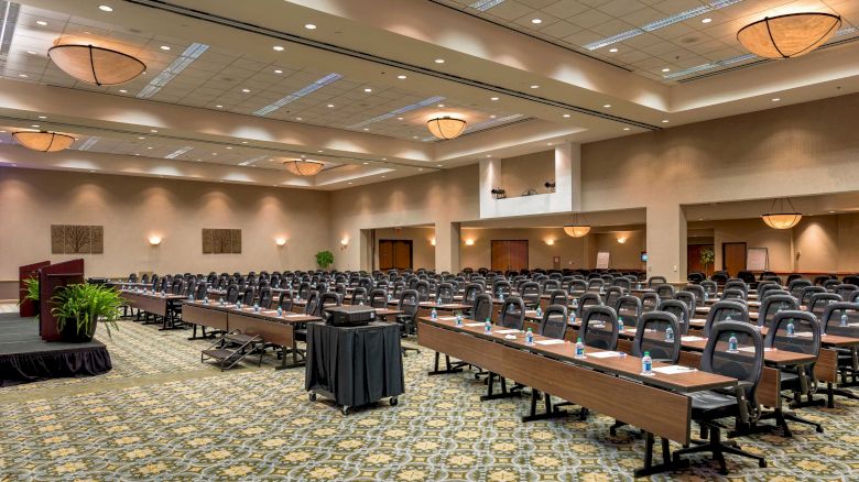 The image shows a large conference room set up with tables, chairs, and a podium on the left side. The room is well-lit with chandeliers.