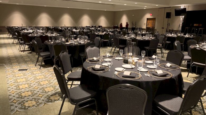 The image shows a large banquet hall set up for an event with round tables covered in black tablecloths, each arranged with place settings.
