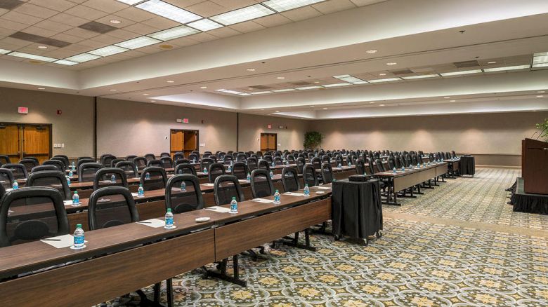 The image shows a large conference room with rows of tables and black chairs. Each table has bottled water and notepads. The room is empty.