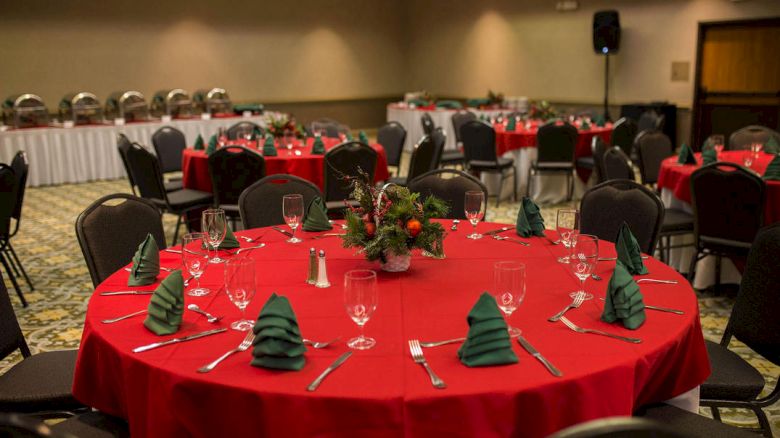The image shows a banquet hall set up for a festive event with tables covered in red tablecloths and green napkins folded like Christmas trees.