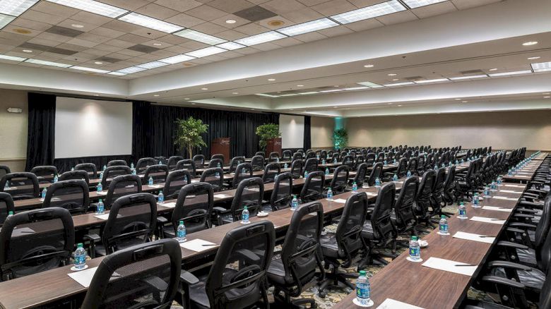 A large conference or meeting room with many chairs, desks, and water bottles, set up for an event or seminar. There are two screens at the front.