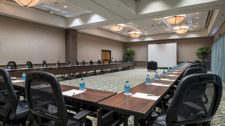 A large conference room with long tables set up in a U-shape, equipped with water bottles, notepads, and chairs; a projector screen at the front.