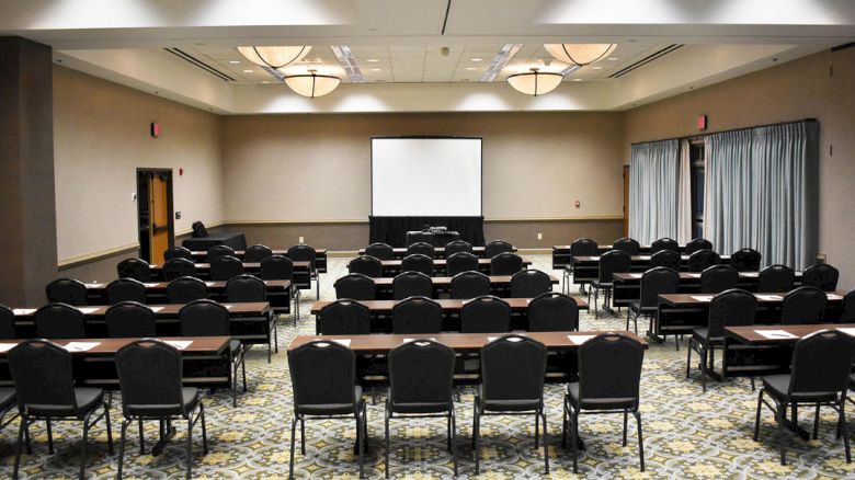 The image shows a conference room with rows of tables and chairs facing a projector screen, ready for a presentation or meeting.