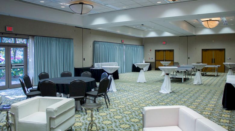 An empty conference room features round tables, cocktail tables, white couches, large windows with drapes, and soft overhead lighting.