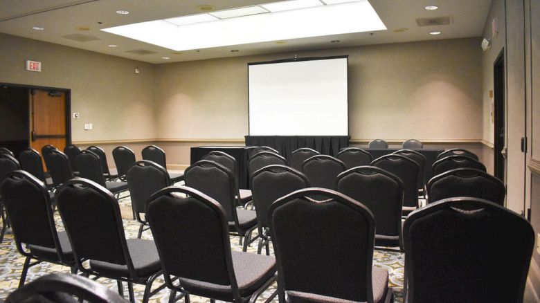 The image shows an empty conference room set up with rows of black chairs facing a projector screen at the front, with overhead lighting.