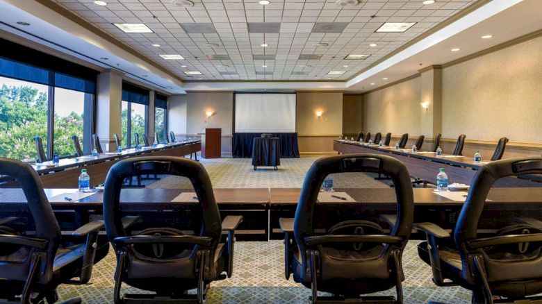 The image shows an empty conference room with long tables arranged in a U-shape, chairs, a projector screen, and water bottles on the tables.
