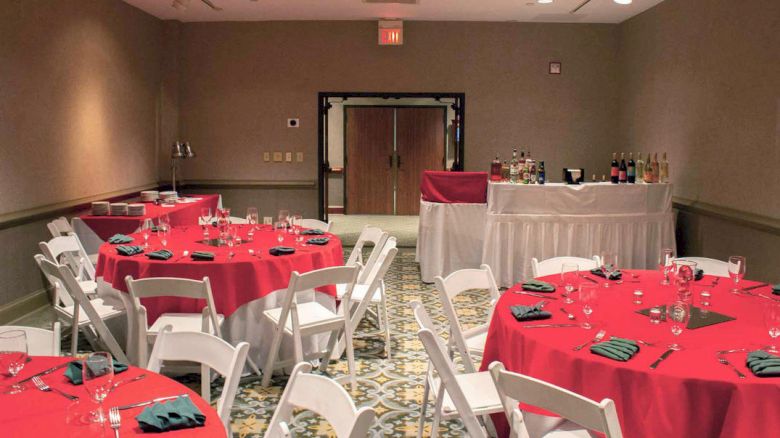 This image shows a banquet room with round tables covered in red tablecloths, white chairs, and a bar setup in the back.