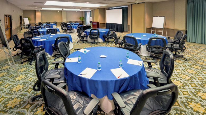This image shows a conference room with round tables, blue tablecloths, black chairs, notepads, water bottles, a projector screen, and flip charts.