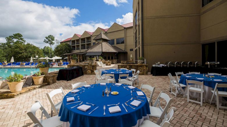 An outdoor banquet setup with tables covered in blue tablecloths and white chairs, alongside a pool and a large building under a partly cloudy sky.
