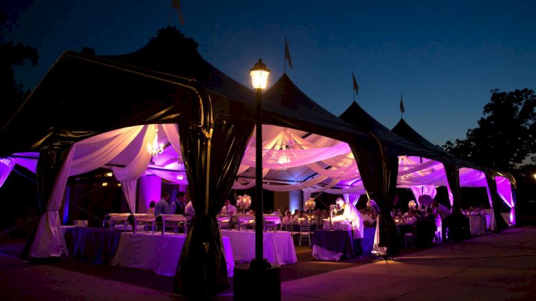 A beautifully lit outdoor event tent at night, adorned with purple lights and draped fabric, possibly for a wedding or special event.