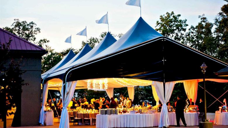 An outdoor event under large, white tents with tables, chairs, and people gathered, illuminated by warm lights, during the evening.