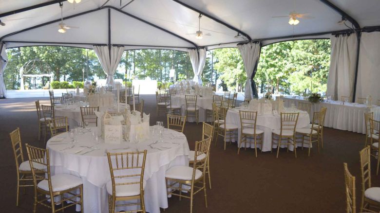A beautifully set up tent with round tables and chairs, likely for an outdoor event or wedding, with greenery visible outside the tent.