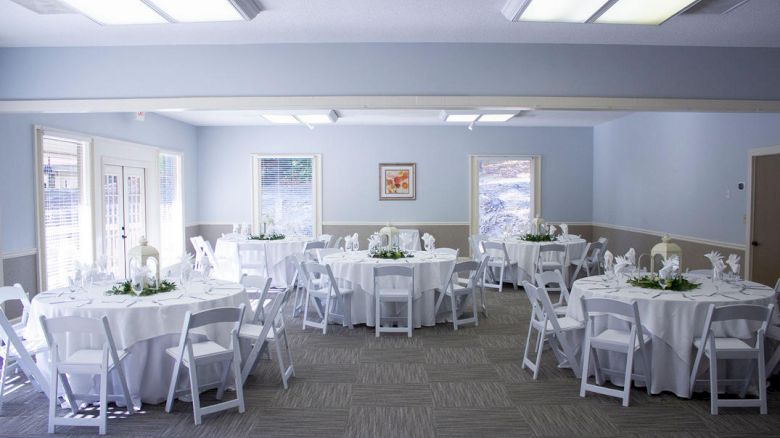 A neatly decorated event room with round tables covered in white tablecloths, white chairs, and centerpieces. The room has a calm, bright ambiance.