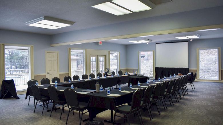The image shows a conference room setup with U-shaped tables, chairs, water bottles, notepads, and a projection screen in a well-lit space.
