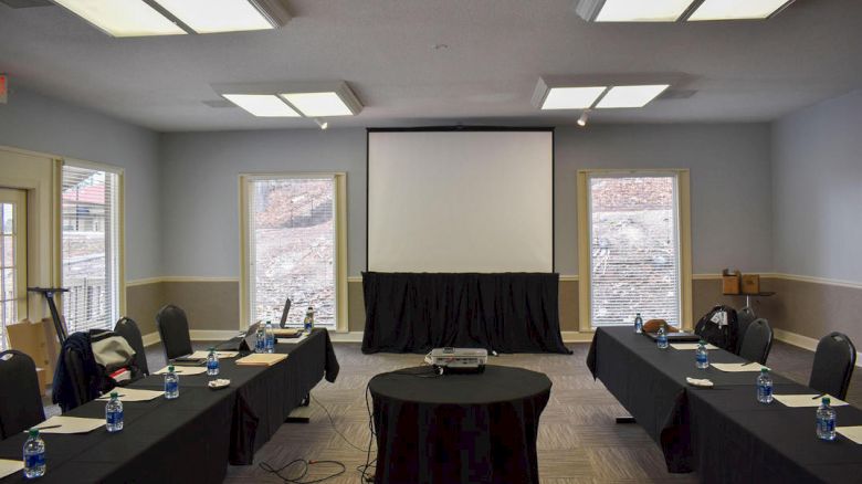 A conference room with a projector screen, U-shaped table setup, chairs, bottled water, and a projector on a round table in the center.