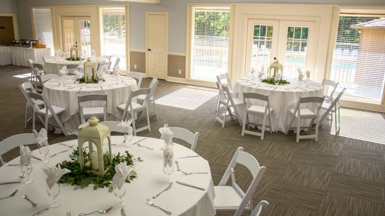 The image shows a decorated event space with round tables set for guests. Each table has white linens, chairs, and a central lantern.