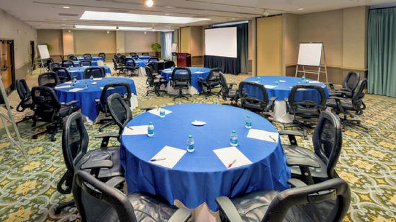 A conference room setup with round tables covered in blue tablecloths, each having chairs, notepads, pens, and water bottles. A projector screen and whiteboards visible.