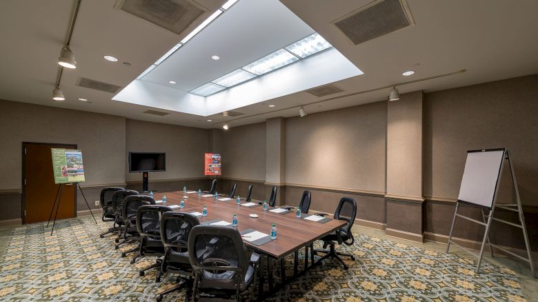 A well-lit conference room with a long table, numerous chairs, a TV screen, flip charts, and whiteboards arranged for a meeting or presentation.