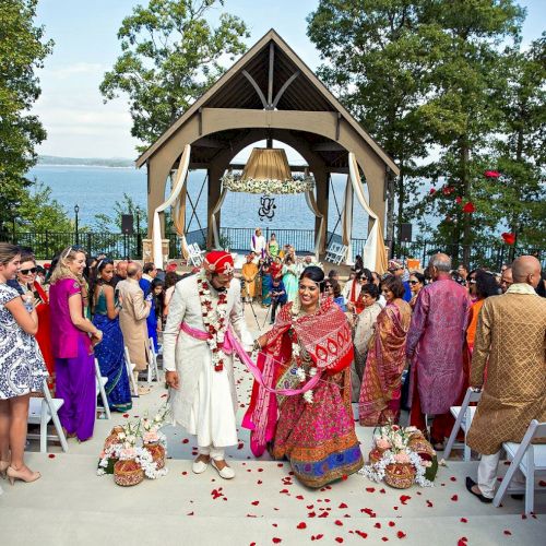 A colorful outdoor wedding ceremony with guests taking photos, an elegant pavilion, and a scenic water view in the background.